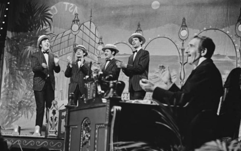 A black and white image showing a barbershop quartet singing on stage, with a man at a desk in the foreground