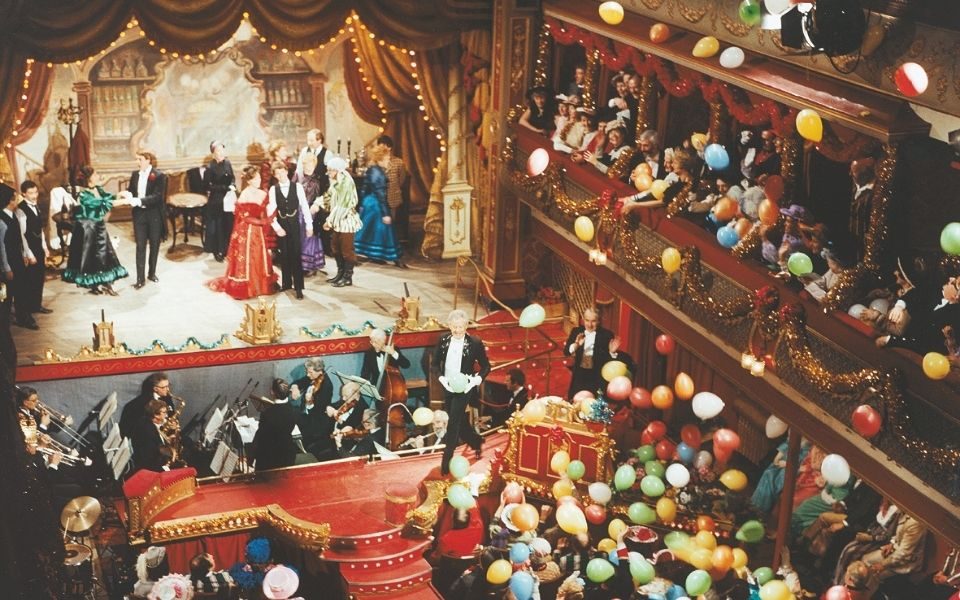 An image showing City Varieties Music Hall in vivid colour, performers and audience members in Victorian attire, as multi-coloured balloons fall from the upper circle