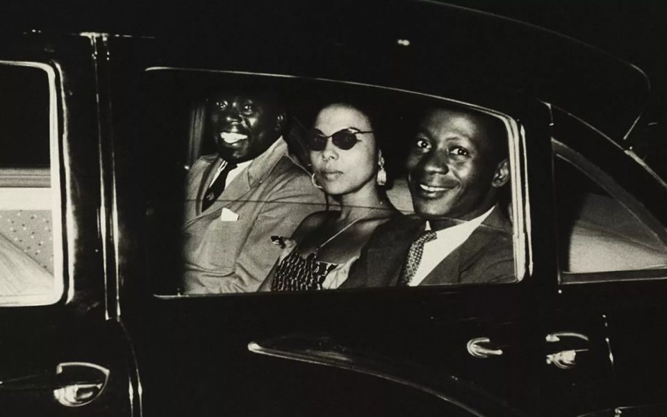 Black and white image. Two smiling men and a woman wearing sunglasses ride in the back seat of a car.