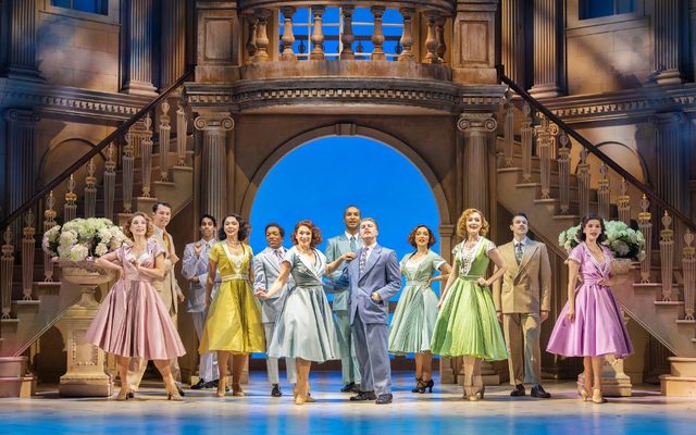 On an elaborate set with a large ornate staircase, a group of actors in pastel coloured suits and dresses stand together in a row.