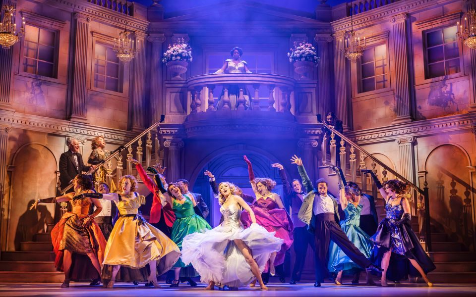 On an elaborate set with a large ornate staircase, a group of actors in colourful dresses and suits perform a choreographed dance, lit by blue haze.