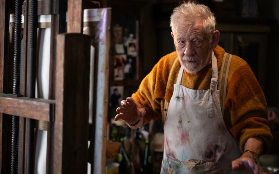 Ian McKellen wearing a fluffy yellow jumper and paint-stained decorators' apron next to a canvas and easel.
