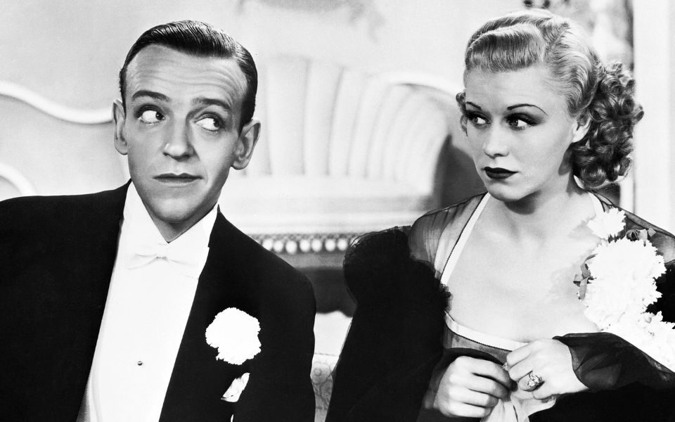 Black and white still. A man in a suit and white bowtie glances across at a woman dressed in a glamorous dress and make-up.