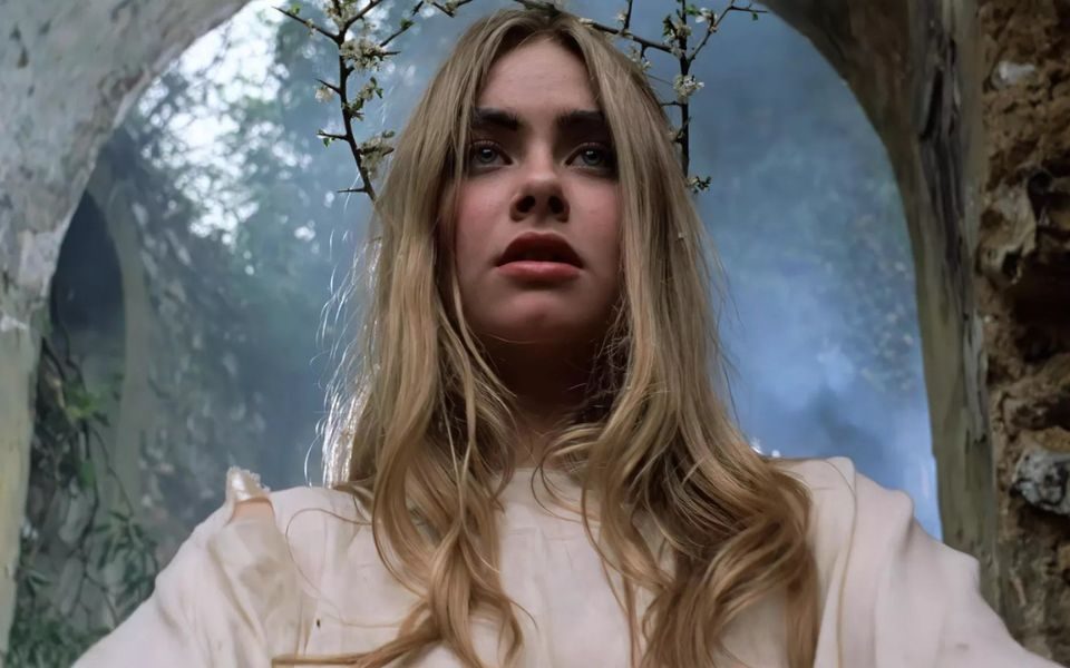A blonde-haired girl in a ripped white dress and a crown of thorns stands beneath a stone arch in the woods.