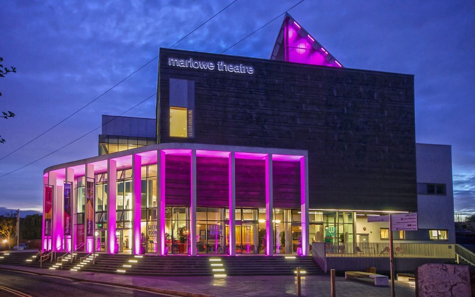 The exterior of The Marlowe Theatre, Canterbury, lit up in purple as the sky gets dark.
