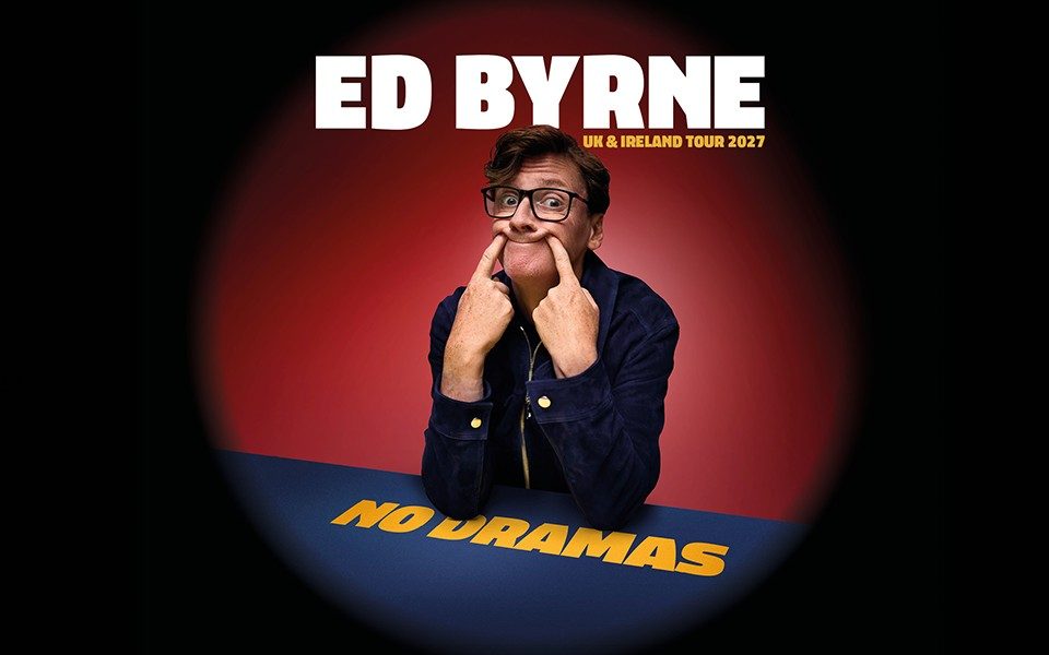 Ed Byrne poses, poking his fingers into his face to form a humorous forced smile. Against a red background, he leans his elbows on a dark blue table. Text reads: Ed Byrne. UK & Ireland Tour 2027. No Dramas.