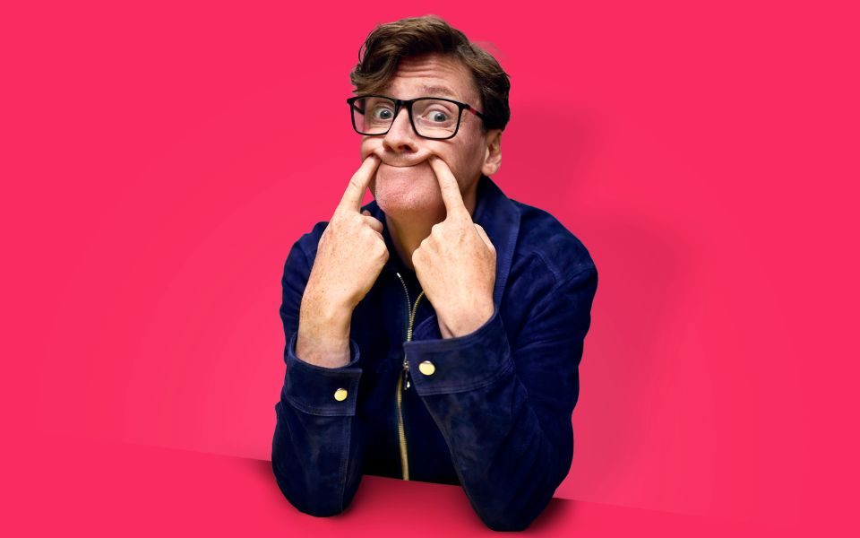 Ed Byrne poses, poking his fingers into his face to form a humorous forced smile. The photo is against a pink background while he leans on a pink table that blends in.