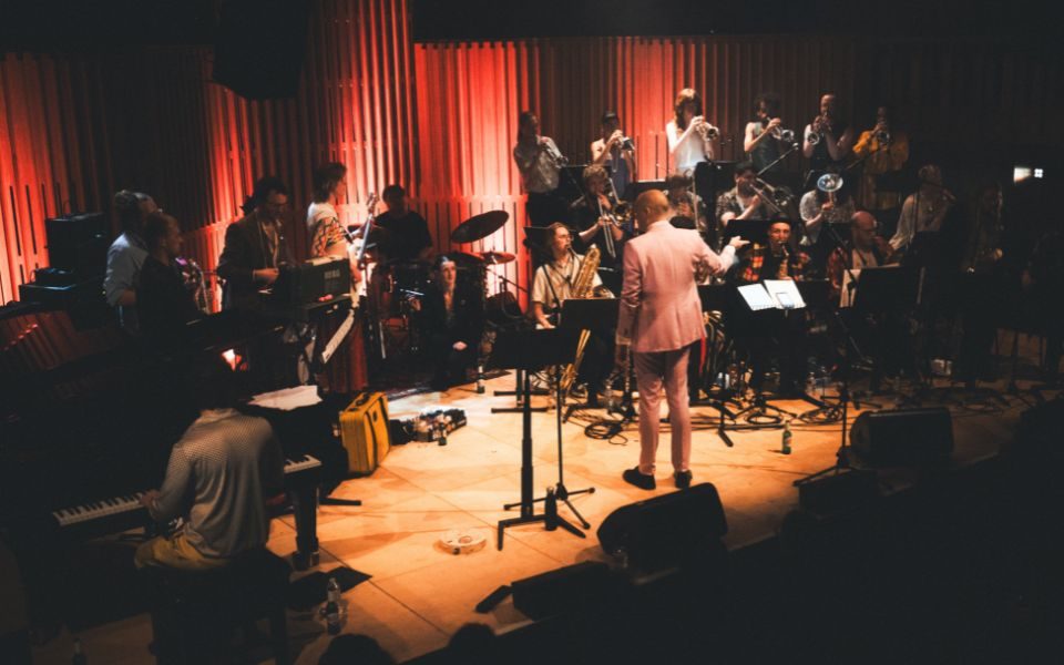 A large collection of musicians in a big band on stage at Howard Assembly Room, being conducted by Fergus Quill in a baby pink suit.