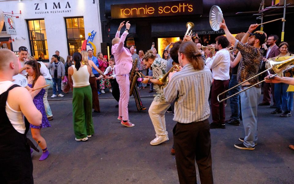 A crowd of musicians in the street outside Ronnie Scott's in London. They play a variety of Big Band instruments, including trombones, trumpets and saxophones. The band leader, Fergus Quill, is leaping in the air dressed in a baby pink suit.