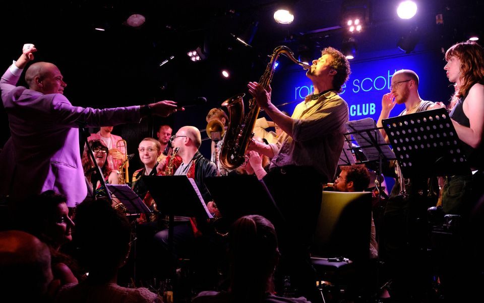 A saxophonist stands up while playing at Ronnie Scott's. Around him sit other members of the band as Fergus Quill, dressed in a baby pink suit, conducts.