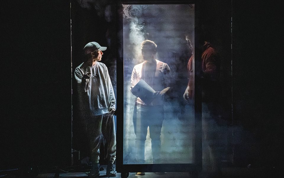Three shadowy figures stand in a row. Two stand behind a rectangular screen looking at the other. Haze surrounds them in the dark.