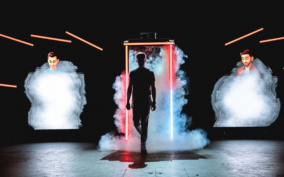 A man silhouetted as he walks out of a smoke-filled frame lit up by blue and red LED lights. Two video game male figures appear on either side out of projected smoke.
