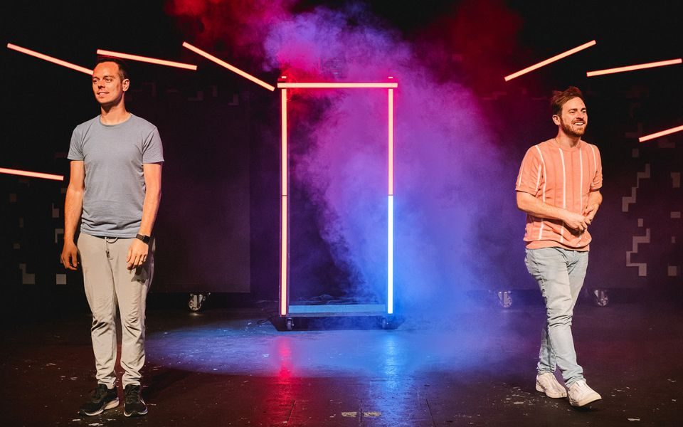 Jay and Joss standing on opposite sides of the stage. Between them is a rectangular frame lit in red and blue with haze emphasising the colours.