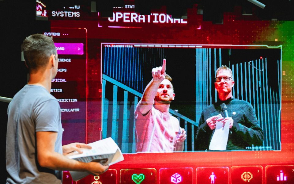 Jay stands holding a script looking upstage at a giant screen with a cut out section. Behind that, Joss points upwards while talking to another man holding a microphone.