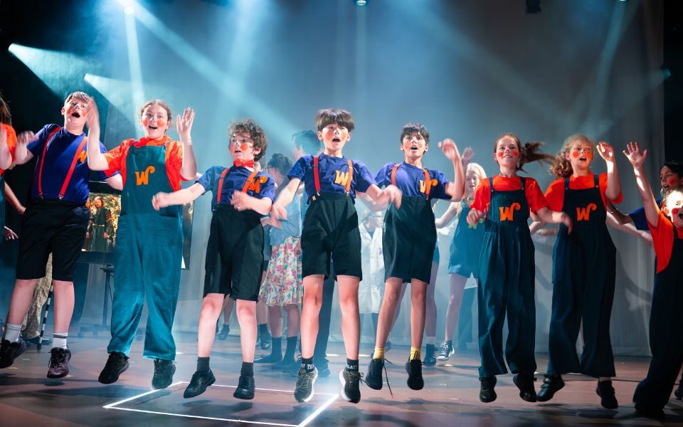 A line of children aged 8-11 are lined up on stage jumping. They are wearing Orange or purple t-shirts and blue shorts or dungarees. They all have orange or purple W's on their tops for Wonka. They are under stage lights and are mid-air in a jump.