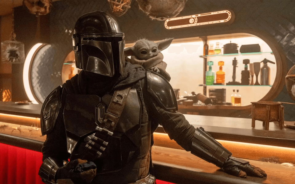 An armoured character with a helmet has a small green creature with long ears resting on his shoulder beside a futuristic bar.