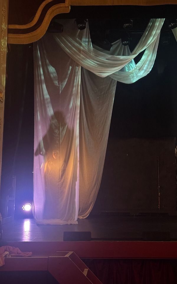 On an empty stage, long fabric is draped dramatically, backlighting creating the silhouette of a figure against it.
