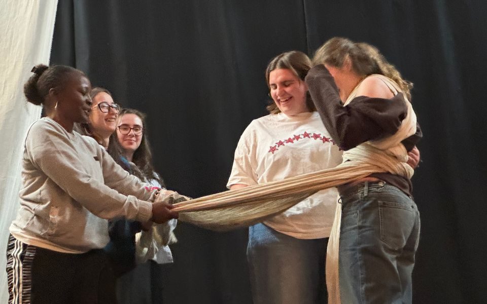 Cast members stand in a group as their choreographer demonstrates, wrapping a length of fabric around a performer's waist.