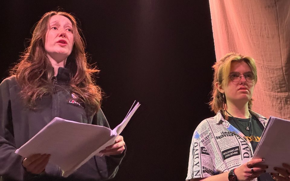 Two cast members speak intensely, holding scripts.
