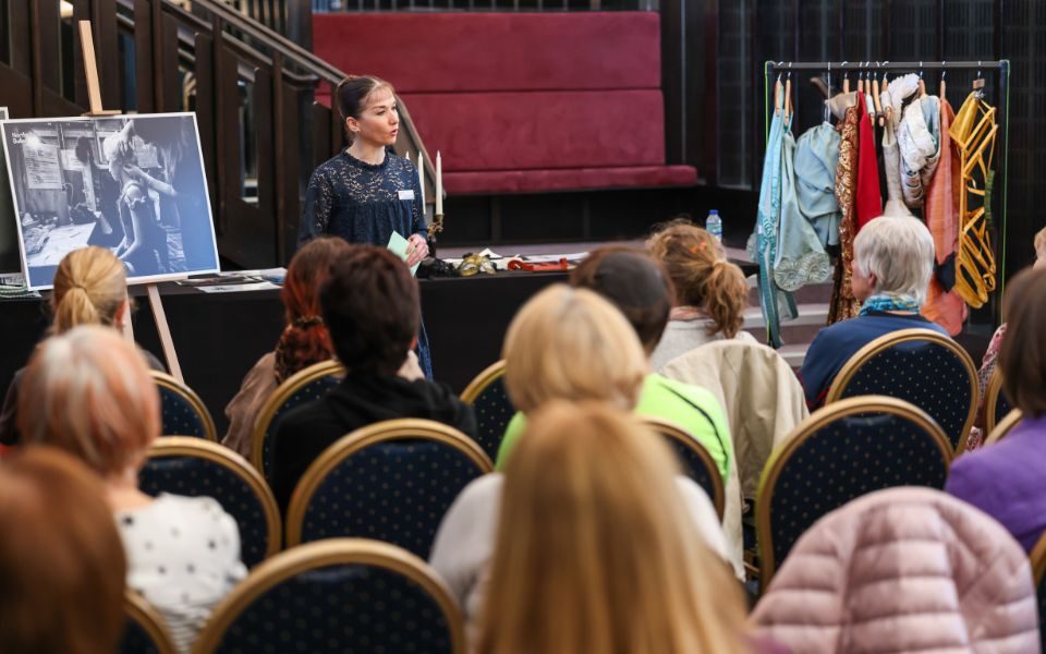 In front of an easel, table and clothing rail of costumes, a woman talks to an audience sat in rows of chairs.