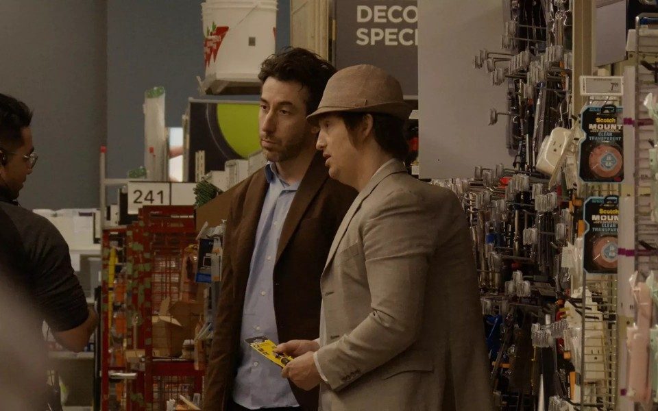 Two men in jackets are in a hardware store, talking candidly to someone off-camera.