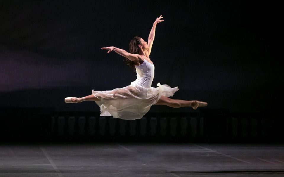 A ballerina jumps in the air with arms raised and legs apart, wearing a flowy white dress on a dark stage.