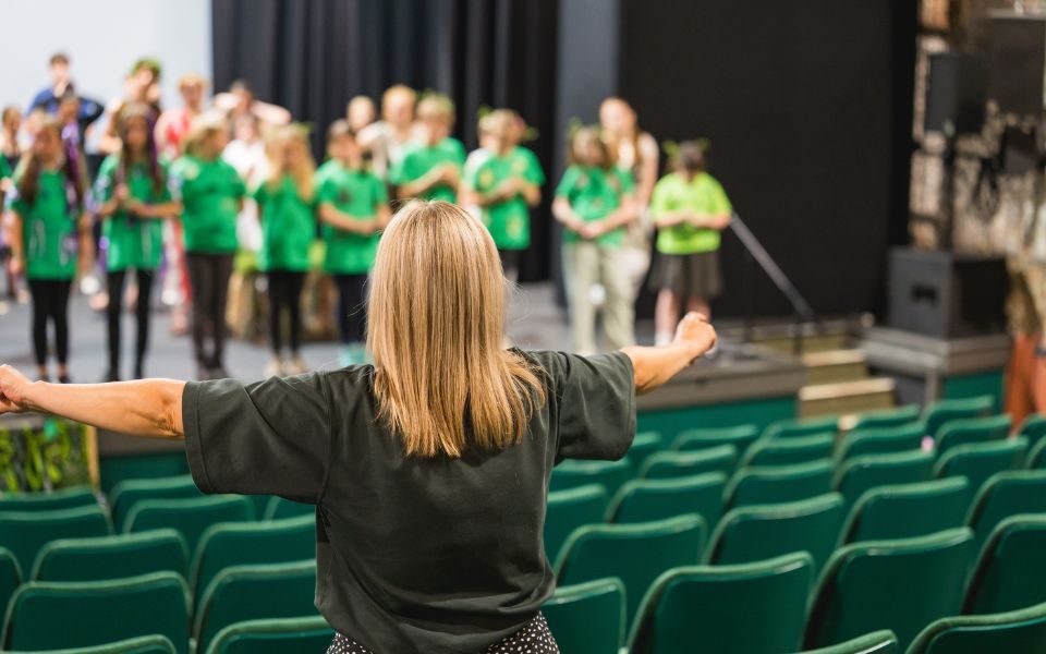 The back of a woman with blonde hair standing with her arms outstretched in the auditorium of Norwich Theatre Royal. On stage are a group of children in matching green tops.