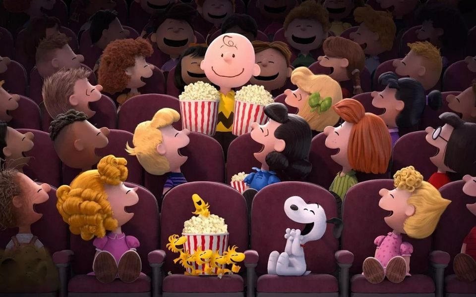 Animated still. Smiley cartoon character Charlie Brown holds two buckets of popcorn in a busy cinema with other children and a white dog, who all face him, laughing.