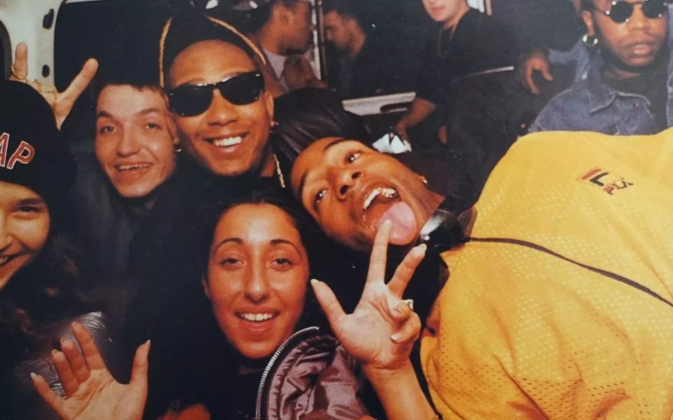 A group of young people in sunglasses, tracksuits and streetwear pose for the camera with hand gestures and one sticks their tongue out.
