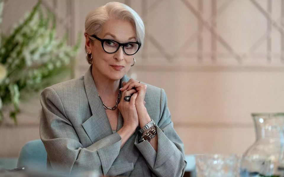 Meryl Streep with short hair, glasses, silver jewellery and a pale grey suit jacket is sat down at a table, hands clasped together.
