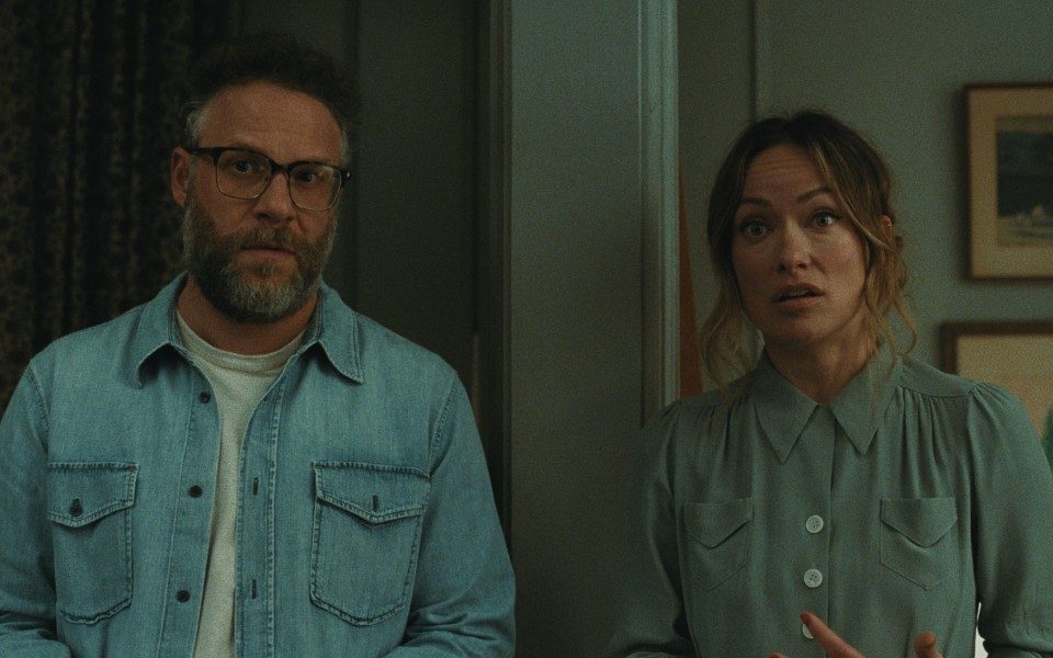 Two people look off-camera with slightly confused expressions, in a room with pale blue walls and framed photos in the background.