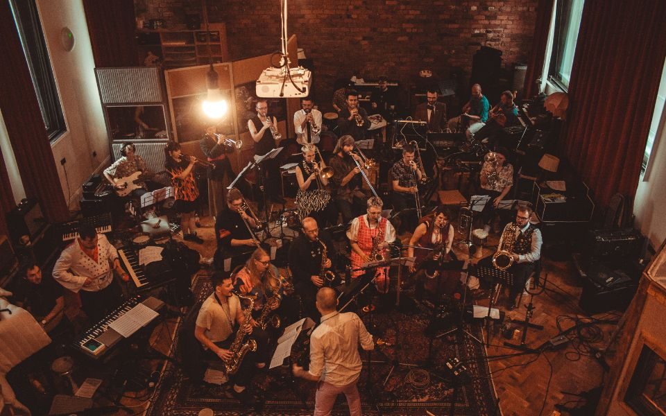 A group of over 30 musicians sitting at chairs with music stands in a recording room for The New Atomic. Fergus Quill stands in front of them conducting.