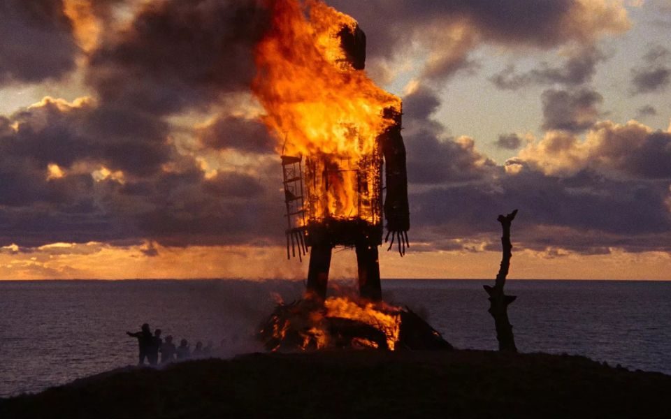The silhouette of a large wicker statue burns on a hill, in front of small figures, a dark sea and cloudy sky.