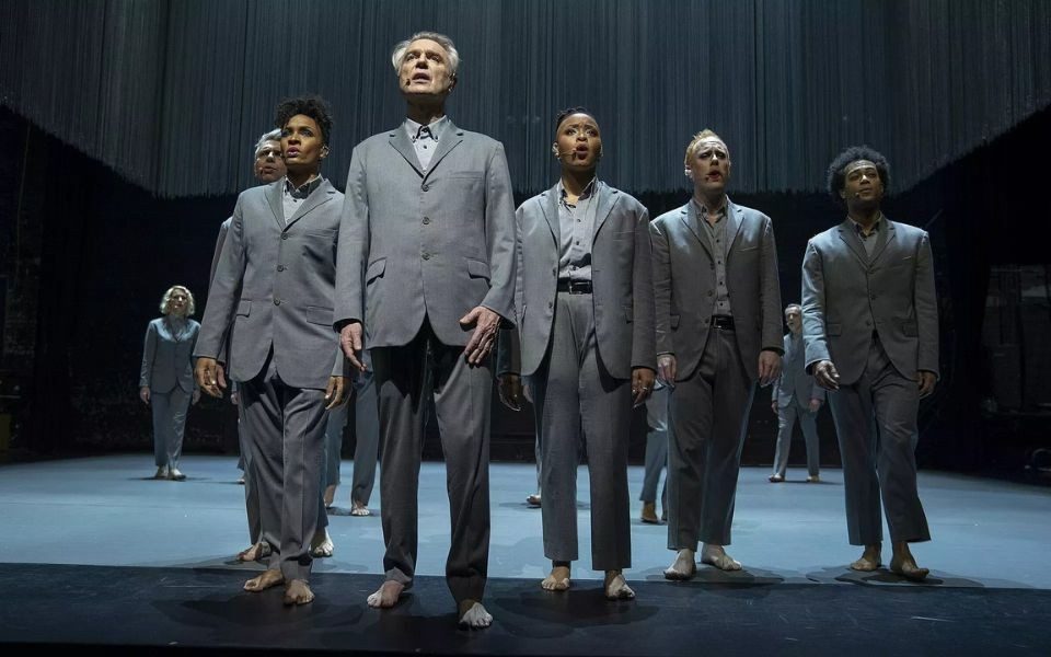 In bare feet and matching grey suits, a group of people stand and sing together on a stark stage under spotlights.