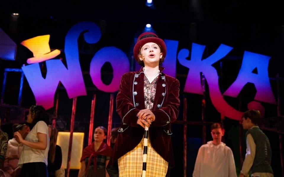 A young person on stage playing Willy Wonka, singing in front of a large Wonka sign