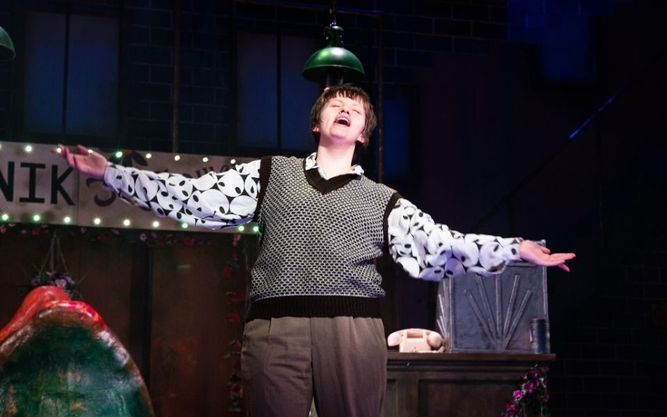 A young person in a sweater vest singing with arms outstretched