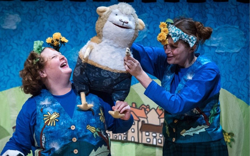 Two people in colourful illustrated waistcoats and flower headbands perform with a guinea pig puppet.