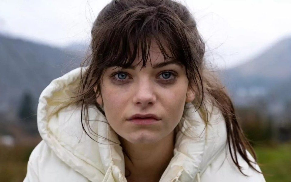 Close-up of a woman's face dramatically looking into the camera, wearing a white puffer coat with windswept hair.