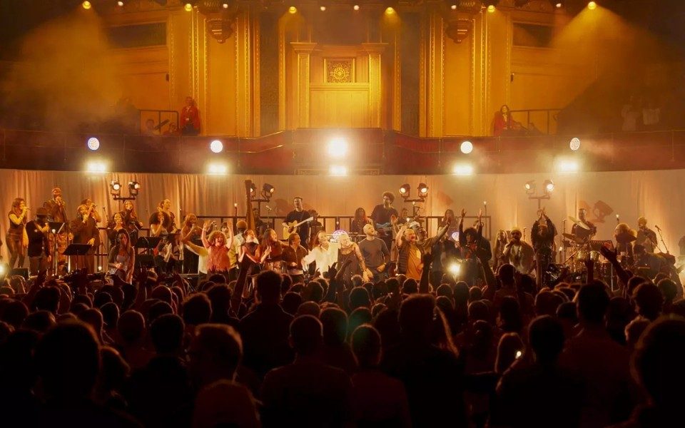 A packed crowd at the Royal Albert Hall with a stage full of dancers and musicians lit by hazy golden lights.