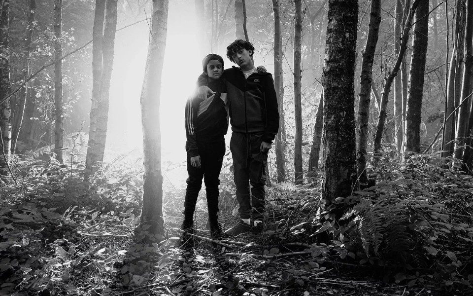 Black and white film still of two children in tracksuits stood in a sun-lit forest, with their arms around each other.