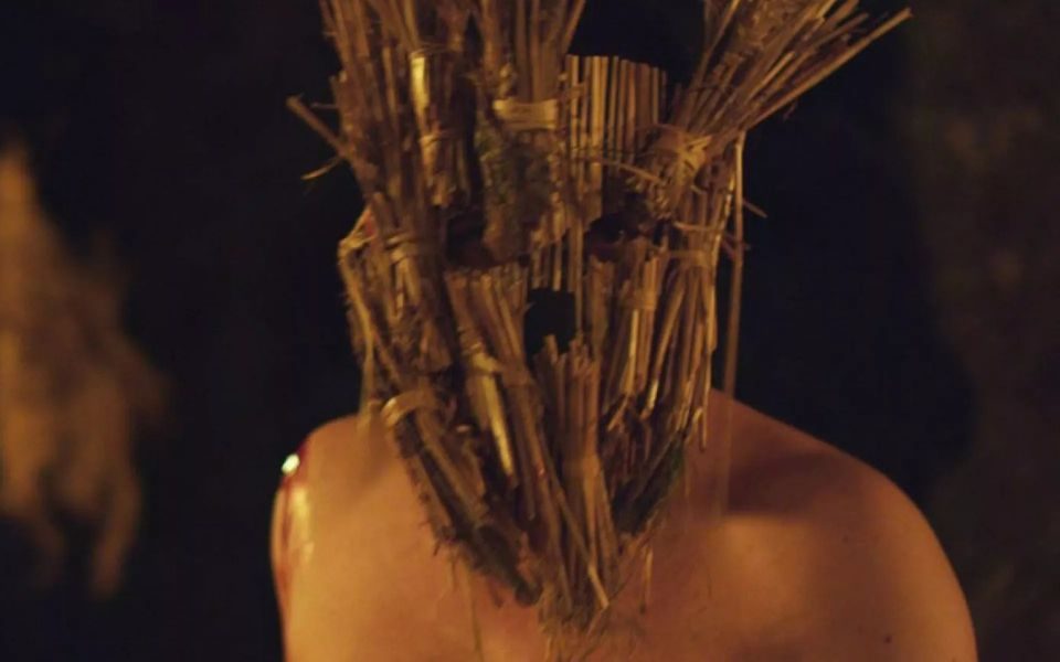 In dark lighting, a man with a bloody shoulder wears a mask of wicker and straw, obscuring his face.