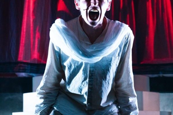 A young man in a tunic is on his knees, screaming. A group of women in flowy dresses are lit by a red glow in the background.