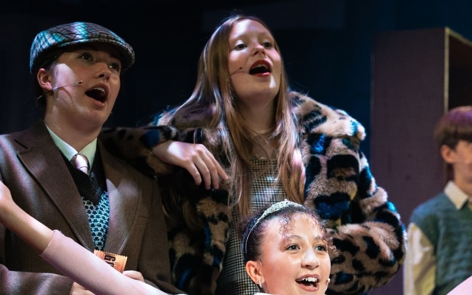 In a huddle with other performers, a young person sings in a fur coat