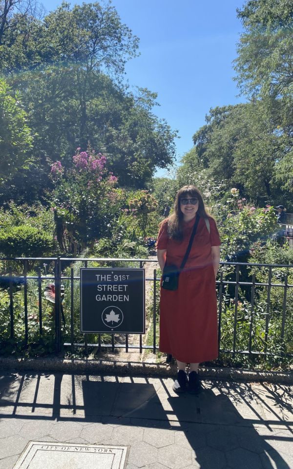 Martha Smiling on a bridge with a sign that reads The 91st Street Garden, with a lavish garden behind her.