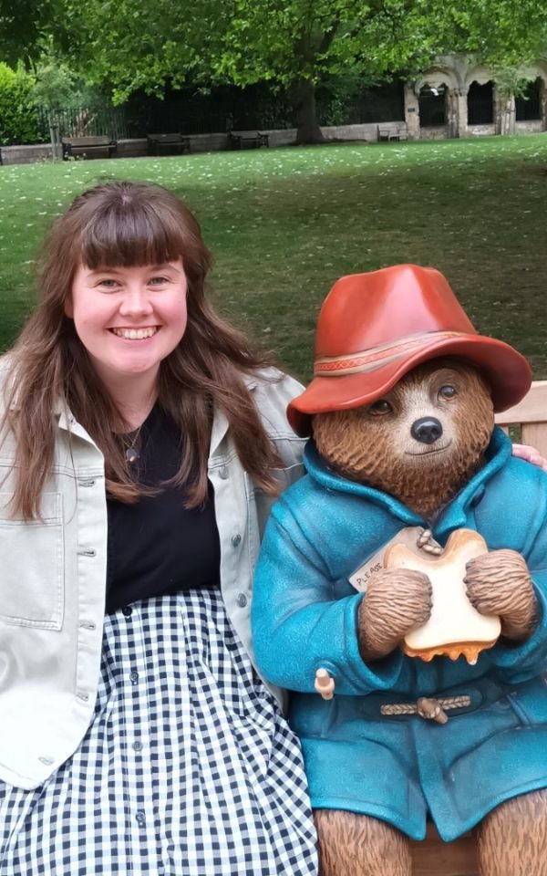 Martha smiling, posing on a bench with a statue of Paddington eating a marmalade sandwich