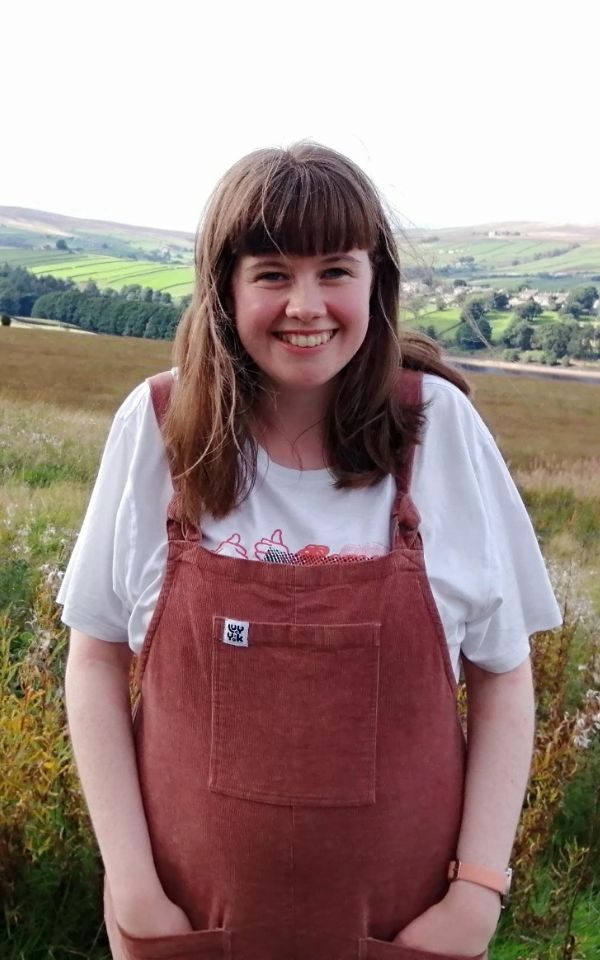 Martha smiling in red dungarees with a country fields in the background.
