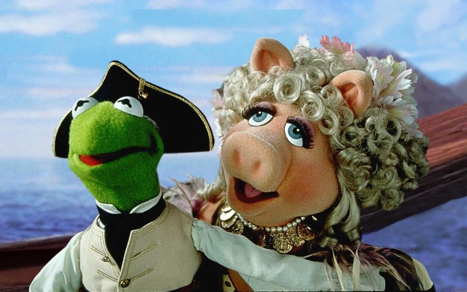Kermit, a green frog puppet, in a pirate hat with Miss Piggy, a pale pink pig puppet with curly blonde hair against a sea backdrop.