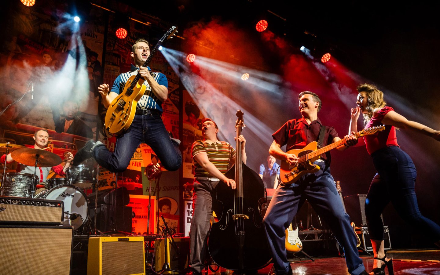 Live production shot of musicians performing on stage, with a guitarist leaping in the air beneath white and red spotlights.