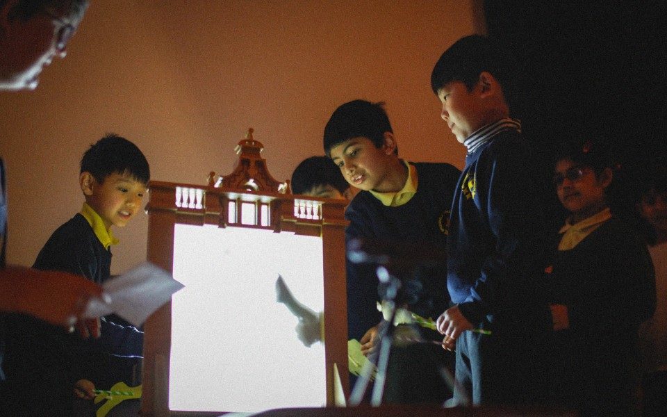 A group of children in school uniforms create shadows using cardboard cutouts behind a light box.