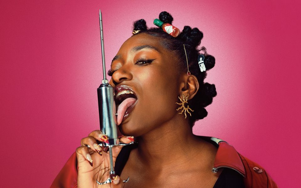 Sophie Duker sticking her tongue out and licking a metallic syringe, wearing fun jewellery and hairclips.